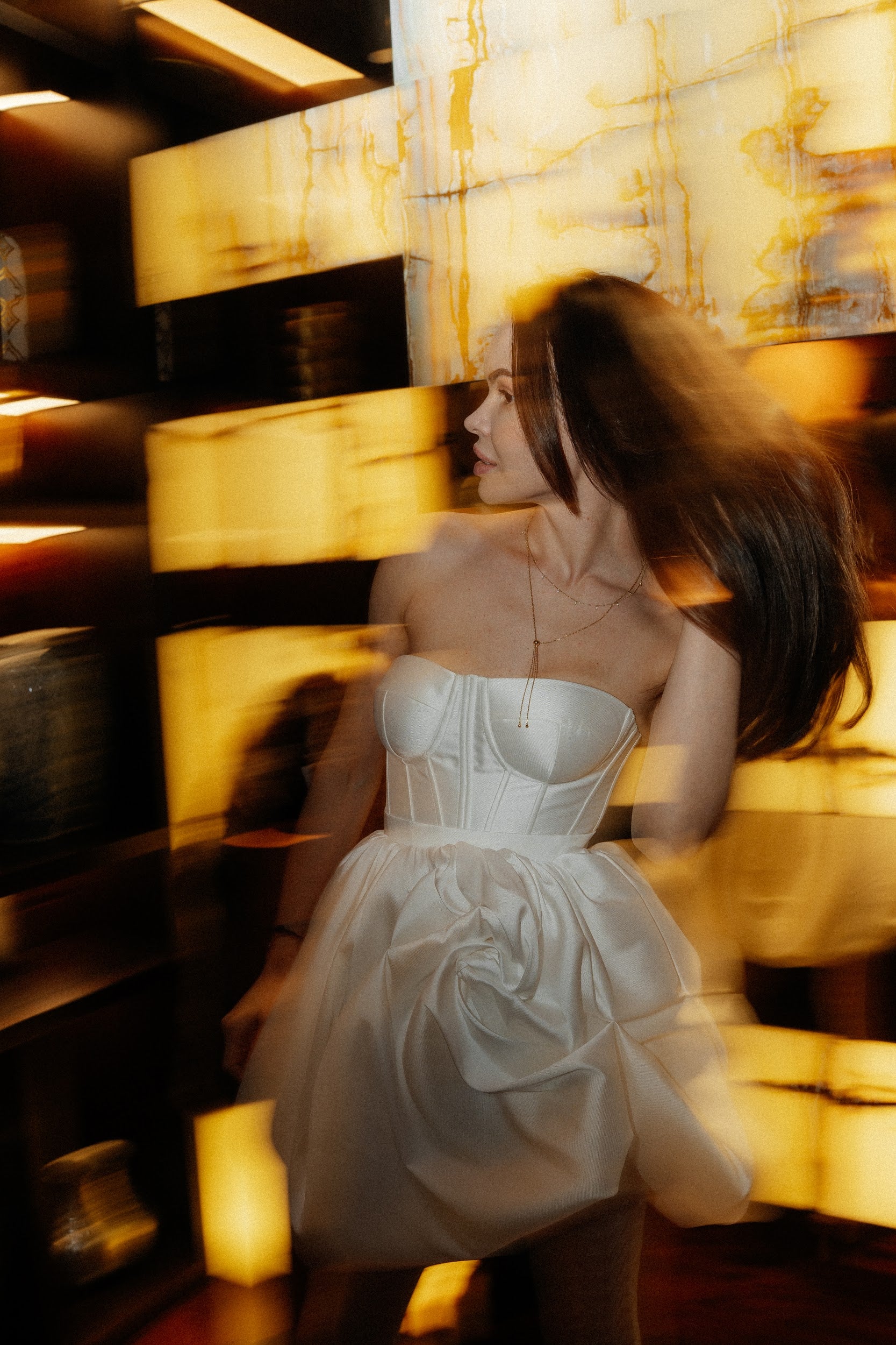 Woman in strapless white dress with a floral detail posing with blurred yellow light panels background